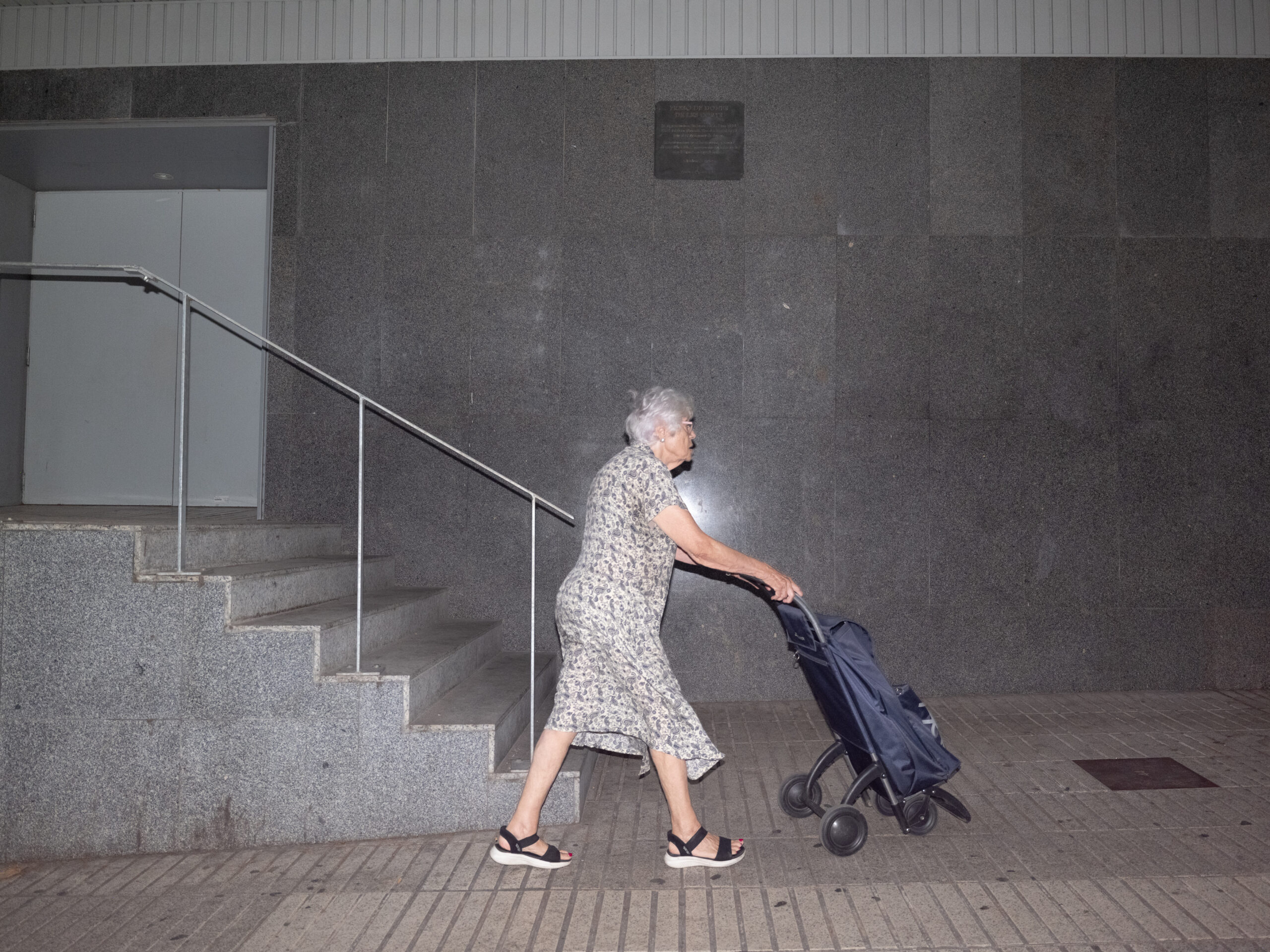 A woman with a shopping trolley passes beneath the Les Corts plaque on the wall of El Corte Inglés, Diagonal, Barcelona, August 2026
